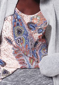 Pestrý top s paisley vzorem v odstínech modré, zelené a oranžové, nošený pod světle šedým kardigánem. Viditelná texturovaná tkanina.