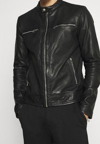 Černá kožená bunda s límcem do stoje, asymetrickými zipovými kapsami a přiléhavým střihem. Hladká textura s lesklým povrchem.