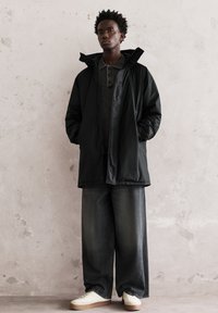 Tânăr îmbrăcat cu o jachetă puffer neagră, blugi largi închiși la culoare și adidași albi, sprijinit de un perete texturat deschis la culoare.