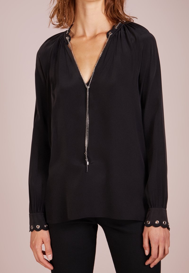 Femme portant un chemisier noir à manches longues avec un col en chaîne argentée et des poignets festonnés à œillets, associé à un pantalon noir.