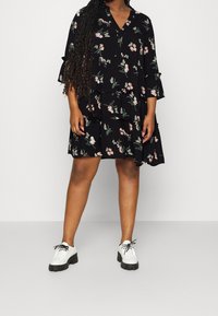 Vestido negro con estampado floral, escote en V, mangas tres cuartos y diseño en capas; combinado con zapatos blancos con suelas gruesas negras.