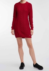 Robe pull en tricot rouge avec un motif en câble, manches longues, col rond et un petit logo en bas. Tissu texturé.