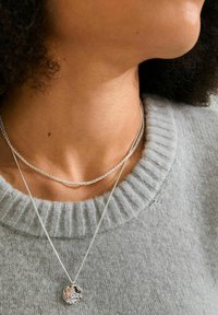 Silvriga halsband lager på lager över en grå, ribbad tröja. Ett halsband har ett texturerat, cirkulärt hänge. Slät yta med fin detaljering.