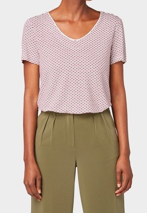 Kobieta ubrana w białą bluzkę z krótkim rękawem i dekoltem w serek z czerwonymi, poziomymi wzorami oraz wysokim stanem oliwkowozielone, plisowane spodnie.