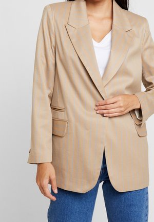 Vrouw die een beige colbert met fijne strepen draagt over een wit topje en een blauwe spijkerbroek, met één hand het colbert gesloten houdend.