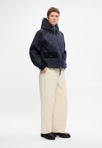 Marineblau gesteppte Jacke mit großer Kapuze, Reißverschluss und schwarzen Stofftaschen. Kombiniert mit cremefarbenen weiten Hosen und klobigen schwarzen Schuhen.