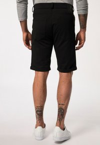 Shorts noirs avec des revers, design slim fit, avec des poches arrière visibles, portés avec des baskets blanches. Tatouages visibles sur les mollets.