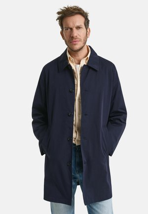 Marineblauwe trenchcoat met een knoopsluiting aan de voorkant, uitstaande kraag en zijzakken. Gedragen over een gestreepte blouse en gecombineerd met lichtblauwe jeans.