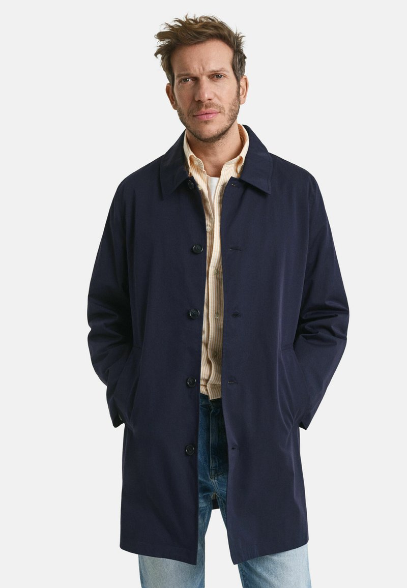 Marineblauwe trenchcoat met een knoopsluiting aan de voorkant, uitstaande kraag en zijzakken. Gedragen over een gestreepte blouse en gecombineerd met lichtblauwe jeans.