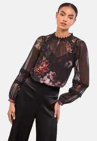 Žena oblečená v priesvitnom čiernom kvetovanom topu s čipkovými detailmi a čiernych nohaviciach s vysokým pásom, pózujúca s rukami položenými pozdĺž tela.