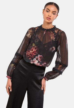 Frau trägt transparente schwarze Bluse mit Blumenmuster und Spitzenakzenten sowie schwarze hoch taillierte Hose, posiert mit seitlich an den Seiten ruhenden Händen.