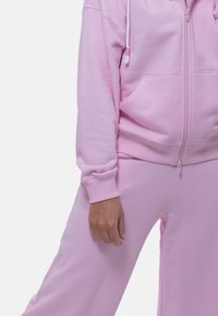 Roze trainingspak van zacht, textuurgevend materiaal. Bevat een hoodie met rits en contrasterende verstelbare koorden, en ontspannen pasvorm broek met zijzakken.