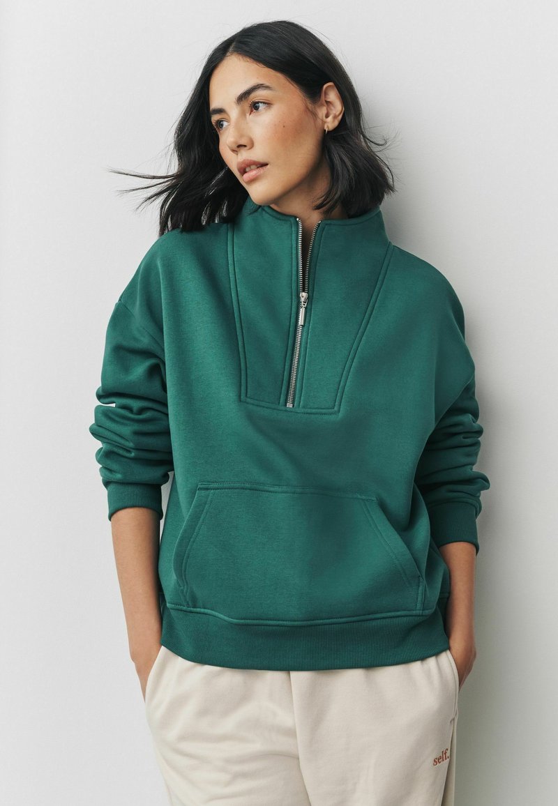 Langærmet grøn sweatshirt med halv-lynlås krave, der har en frontlomme og ribbede manchetter, lavet af blødt stof.