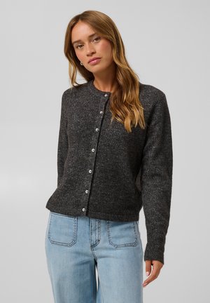Kvinde med langt lystbrunt hår iført en mørkegrå knapcardigan og lyseblå jeans mod en ensfarvet baggrund.