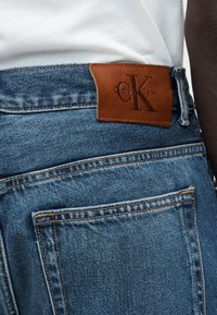 Κοντινό πλάνο της πίσω τσέπης μπλε τζιν με καφέ δερμάτινη ετικέτα Calvin Klein στη μέση, σε συνδυασμό με λευκό πουκάμισο.