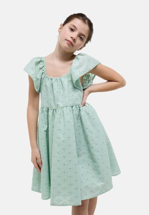 Jeune fille portant une robe en broderie anglaise vert clair à manches courtes avec des épaules à volants, debout avec une main sur la hanche devant un fond uni.