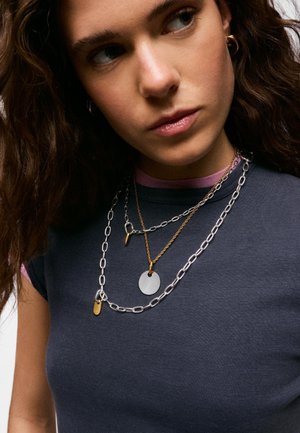 Joven mujer con el cabello castaño ondulado lleva collares de cadenas en capas de plata y oro sobre una camiseta gris oscuro con ribete morado.