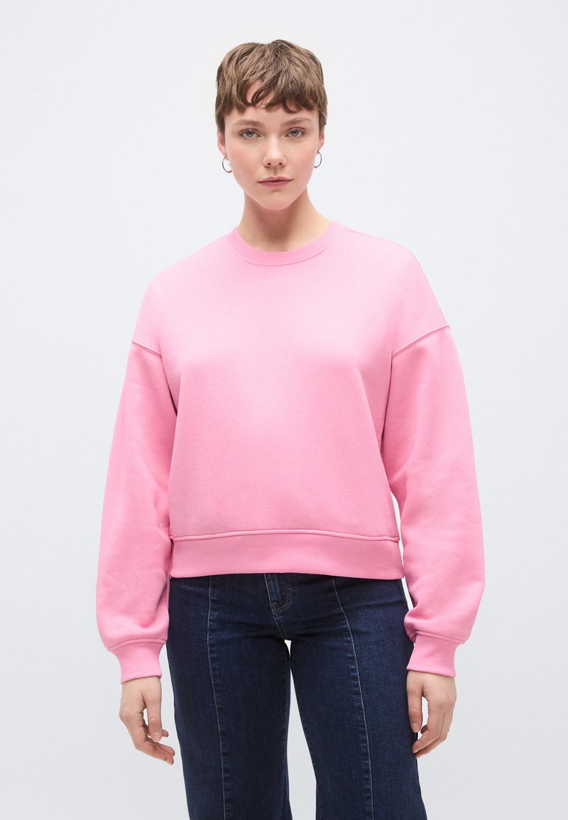 Ροζ φούτερ με στρογγυλή λαιμόκοψη, χαλαρούς ώμους και μανσέτες με σχέδιο ριγκ. Απαλή υφή και άνετη εφαρμογή, συνδυασμένο με σκούρο μπλε τζιν.