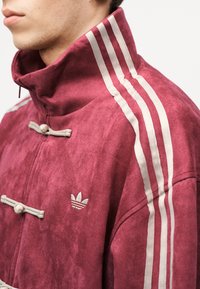 Άτομο που φοράει μπουφάν Adidas από σουέτ σε μπορντό χρώμα με λευκές τρεις ρίγες στον ώμο και κεντημένο λογότυπο στο στήθος, με ορατό ψηλό γιακά.