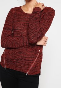 Pull en tricot rouge avec un motif texturé, manches longues et accents de sangle en métal argenté de chaque côté ; design ajusté avec un ourlet en côtes.