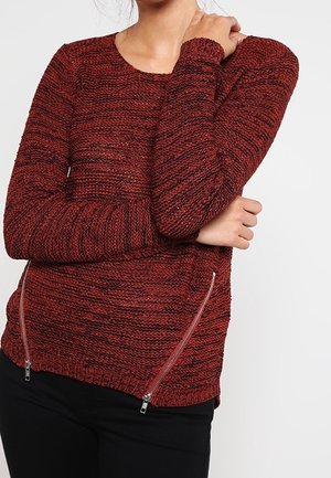 Pull en tricot rouge avec un motif texturé, manches longues et accents de sangle en métal argenté de chaque côté ; design ajusté avec un ourlet en côtes.