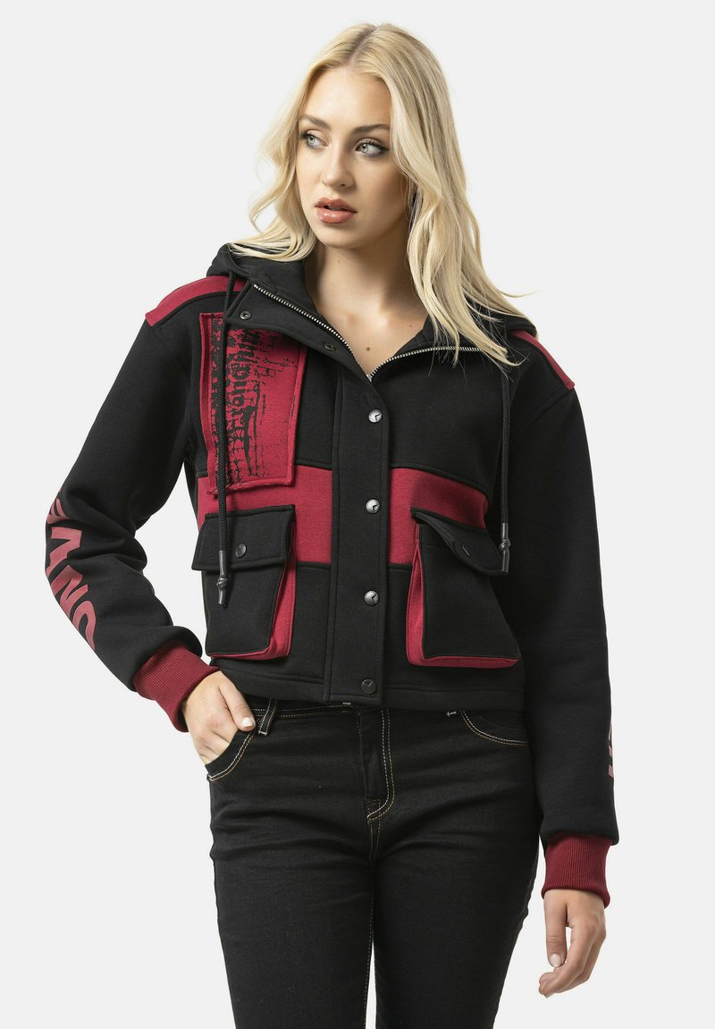 Zwarte en bordeauxrode cropped hoodie met ritssluiting aan de voorkant, twee grote voorzakken en rode accenten. Voorzien van een capuchon met trekkoord en opvallende typografie op de mouw.