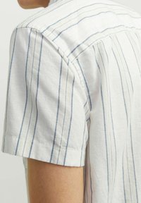 Camisa de manga corta en tela blanca ligera con rayas verticales azules. Presenta un cuello clásico y un dobladillo curvado.