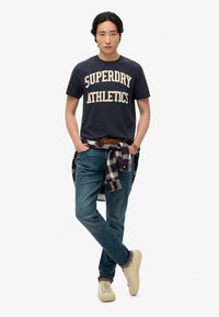 Camiseta navy con "SUPERDRY ATHLETICS" impreso en crema, combinada con jeans azules. Una camisa a cuadros atada a la cintura y zapatillas crema.