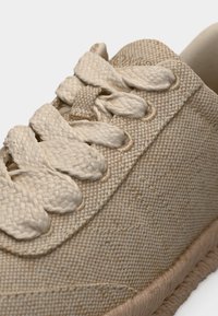 Κοντινό πλάνο αθλητικού παπουτσιού από μπεζ υφαντό ύφασμα με χοντρές πλεγμένες κορδόνια και υφασμένη σόλα σε λευκό φόντο.