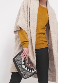 Beige textuur sjaal over een mosterdkleurige trui, gecombineerd met een zwart-wit clutch met patronen, gekenmerkt door geometrische ontwerpen en kleurrijke accenten.