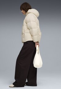 Giubbotto imbottito beige con cappuccio, caratterizzato da un design oversize. Abbinato a pantaloni larghi scuri e a una piccola borsa bianca. Look semplice e moderno.