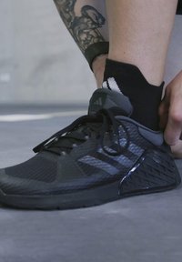 Zapatilla deportiva negra con una parte superior de malla texturizada, logo de tres franjas y acentos laterales con patrón de diamantes. Se están atando los cordones.