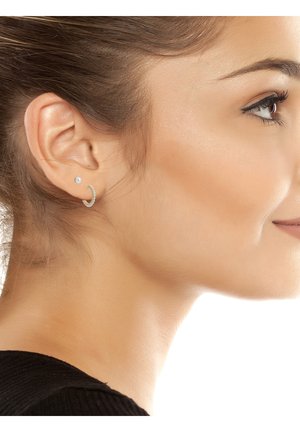 Prawy profil kobiety z czystą skórą, subtelnym makijażem, małym perłowym kolczykiem w sztyfcie oraz srebrną obrączką w uchu.