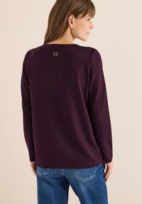 Langeærmet lilla top med rund halsudskæring og et lille logo på ryggen. Lavet af blødt stof, parret med blå denimjeans.