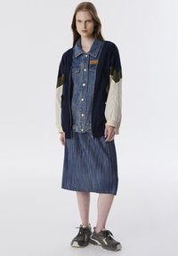 Denimjacket met marineblauwe stof, beige mouwen en een logo-patch. Draag het over een lange, gestreepte denimschoot en grijze sneakers.