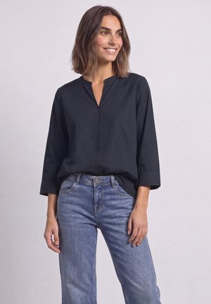 Femme aux cheveux bruns mi-longs souriante, portant un chemisier noir boutonné à manches trois-quarts et un jean bleu, debout devant un fond uni.
