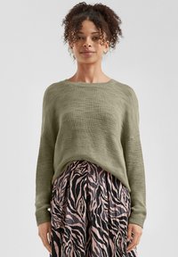 Olivengrüner Strickpullover mit lockerer Passform und gerippten Bündchen, kombiniert mit einem Faltenrock im Schwarz-Pink-Zebra-Muster.