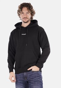 Felpa nera in cotone con tasca frontale e stringhe. Presenta la scritta "PROPAGANDA" stampata in bianco sul petto. Indossata con jeans blu.