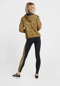 Geel en zwart luipaardprint hoodiejack, gecombineerd met zwarte leggings met een luipaardprint zijkantstreep, gedragen met zwarte sneakers.
