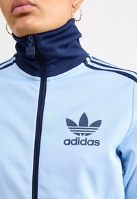 Detailní záběr osoby oblečené v světle modré mikině Adidas se zipem, tmavě modrým límcem a logem, a zkroucenými zlatými náušnicemi ve tvaru kruhu.