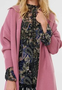 Femme portant un long cardigan rose sur une robe foncée à motifs fleuris et cachemire, tenant le col d'une main, montrée des épaules aux genoux.