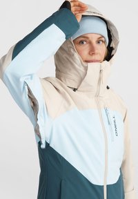 Veste de ski légère et imperméable en beige, bleu clair et sarcelle. Elle dispose d'un col haut, d'une capuche ajustable et de poches zippées.
