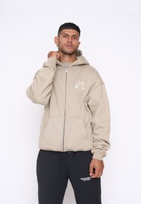 Beige zip-up hoodie med en stor hætte, kængurulommer og hvidt logo detalje på brystet. Parret med sorte sweatpants med hvid tekst.