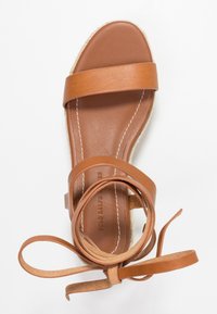 Brun läder sandal med två breda remmar, en ankelsnodd och en texturerad espadrillesula med vit söm.