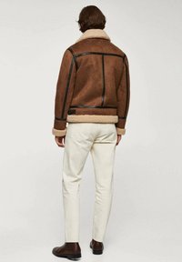 Man in een bruin schapenvachtjack met zwarte accenten, off-white broek en donkerbruine laarzen, staande tegen een effen lichte achtergrond.