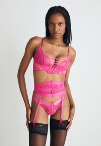 Rosa undertøysett med blonde- og meshdetaljer, som inkluderer en bralette med stropper, et høyhåndt strømpebånd og svarte thigh-high strømper.