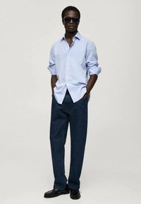 Cămașă bleu deschis cu nasturi, pantaloni din denim închis, pantofi negri și ochelari de soare. Cămașa are mânecile răsfrânte; materialul pare neted și respirabil.
