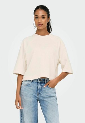 Junge Frau mit Creolen, die ein lockeres weißes, kurzes T-Shirt und hellblaue, hoch taillierte Jeans trägt, mit einer Hand in der Tasche.