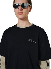 Personne portant des lunettes de soleil argentées réfléchissantes et un T-shirt noir par-dessus une chemise beige à manches longues, avec « HALO » et des chiffres imprimés sur la poitrine.
