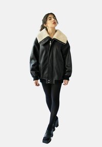 Svart läderjacka med en stor krämfärgad shearlingkrage, framdragkedja och ribbade muddar. Bärs över svarta strumpbyxor och kraftiga klackar.